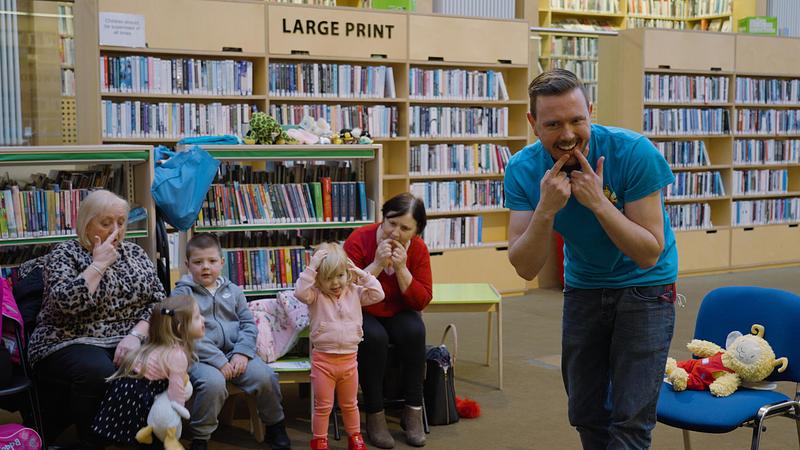 A man in a blue Bookbug t-shirt gesturing to library guests and encouraging them to smile